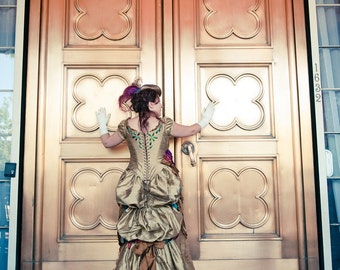 May include: A woman in a gold and brown dress with a green and blue floral design stands in front of a pair of large, ornate, gold doors. The doors have a clover-shaped design.