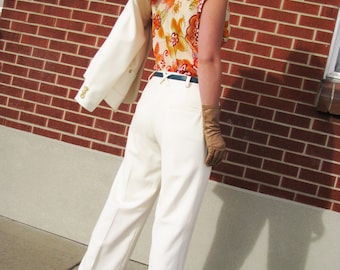 May include: A woman wearing a white blazer, a floral print top, white trousers, and brown leather gloves. The woman is standing in front of a brick wall.