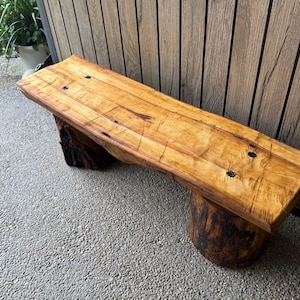 May include: A wooden bench featuring a thick, light brown live-edge slab seat with visible wood grain and dark knots. The seat is supported by two cylindrical, dark brown log bases. The surface of the wood shows natural textures and markings.