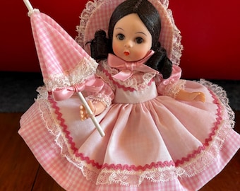 May include: A doll with dark hair wearing a pink gingham dress with white lace trim and a matching bonnet. The doll holds a small pink gingham parasol with lace edges and a pink ribbon bow. The dress features a pink zigzag trim detail.