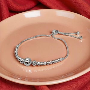May include: A silver adjustable bracelet with a sliding bead closure. The bracelet features small, round beads and a larger central bead. The bracelet is displayed on a pink plate against a red background.