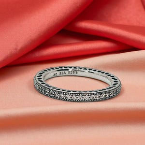 May include: A silver ring with a continuous row of clear gemstones. The ring has a detailed design around the band and is set against a backdrop of pink and red fabric.
