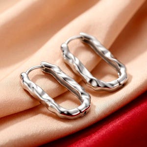 May include: A pair of silver-toned hoop earrings featuring an elongated, oval shape with a distinctively wavy, textured surface. The earrings are designed with a hinged closure mechanism, showcasing a polished metallic finish that reflects light.