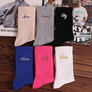 Può includere: Una collezione di calzini ricamati personalizzati in vari colori, tra cui beige, grigio, nero, blu, rosa e bianco. Ogni calzino presenta un nome diverso ricamato in un colore a contrasto. Il testo "Personalized Embroidered Socks" è visibile.