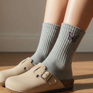 May include: A pair of gray ribbed socks with a purple embroidered butterfly design. The socks are worn with beige suede clogs with a buckle closure. The clogs have a cork sole and are on a wooden floor.