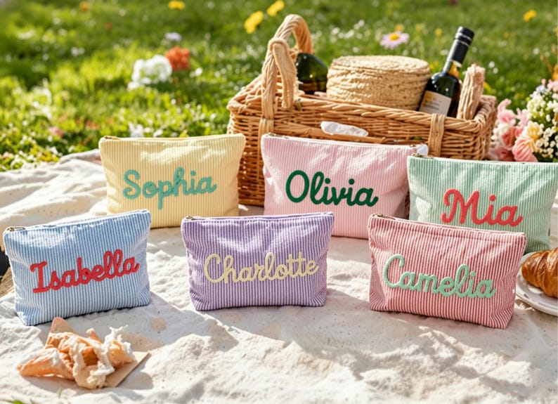 May include: A collection of personalized cosmetic bags in various colors, including yellow, pink, blue, purple, and red, each embroidered with a different name. The bags are striped and arranged on a picnic blanket with a basket and wine bottle in the background.