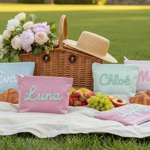 May include: A picnic scene with a wicker basket, straw hat, and several striped cosmetic bags with names embroidered on them. The bags are in various colors, including pink, blue, and mint green. Fresh fruit and croissants are also present.