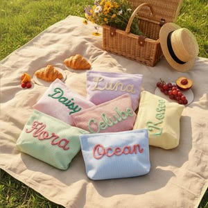 May include: A picnic scene with a woven basket, straw hat, and a beige blanket. Six corduroy pouches in pastel colors are arranged on the blanket, each embroidered with a name: "Daisy", "Flora", "Luna", "Celube", "Ros&eacute;e", and "Ocean".