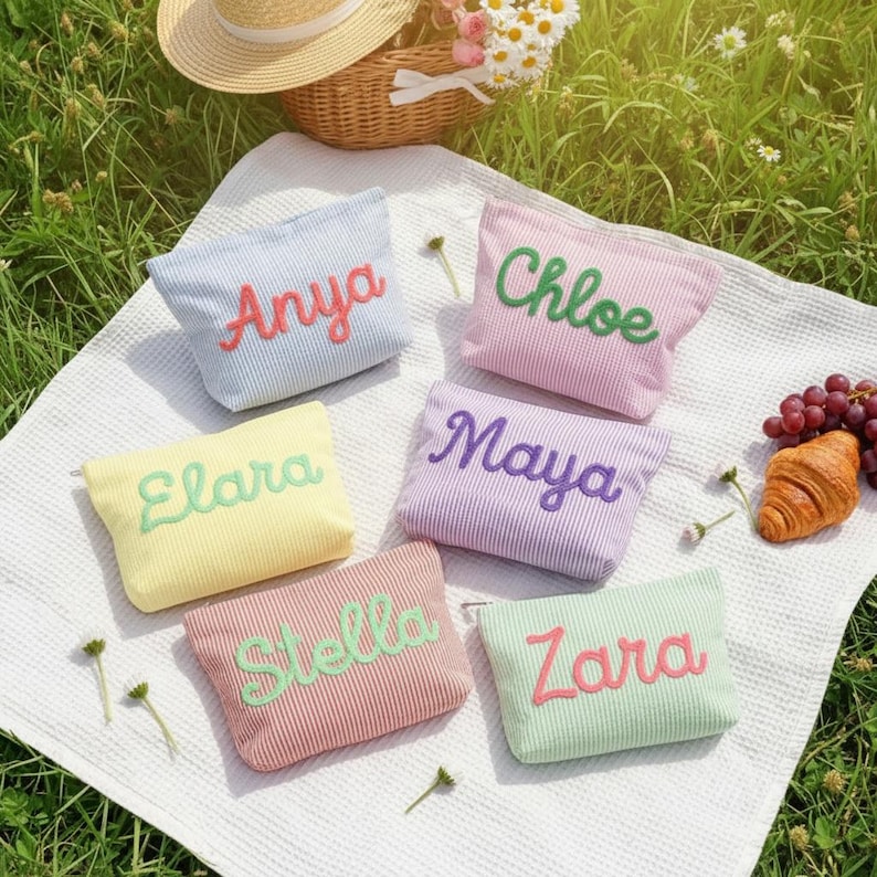 May include: Six pastel-colored, striped cosmetic bags with embroidered names: Anya, Chloe, Elara, Maya, Stella, and Zara. The bags are arranged on a white blanket, with a picnic basket, hat, grapes, and croissant in the background.