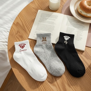Könnte beinhalten: Drei Paar Knöchelsocken in Weiß, Grau und Schwarz, jeweils mit einem gestickten Design und Namen. Die weiße Socke hat ein rotes Emblem und den Namen "Emma". Die graue Socke hat ein braunes Emblem und den Namen "Sofia". Die schwarze Socke hat ein weißes Emblem und den Namen "Lily".