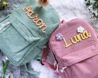 Könnte beinhalten: Zwei personalisierte Cord-Rucksäcke, einer grün mit dem Namen "Aaron" in Gelb gestickt und der andere rosa mit dem Namen "Luna" in Gelb gestickt. Beide Rucksäcke haben eine kleine Tasche auf der Vorderseite und sind mit gehäkelten Blumen verziert.
