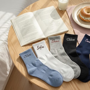 Puede incluir: Cinco pares de calcetines personalizados de varios colores, cada uno bordado con un nombre: Chloe, Sofia, Emma, Chloe y Sarah. Los calcetines están colocados sobre una mesa de madera con un libro, un bagel y una vela.