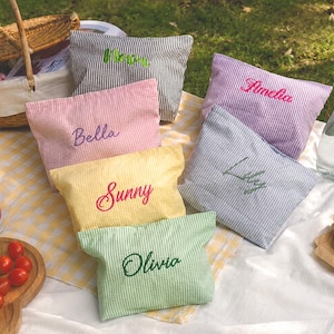 Puede incluir: Una escena de picnic con bolsas de seersucker personalizadas en varios colores, cada una bordada con un nombre. Las bolsas están dispuestas sobre una manta de picnic con comida y bebidas, incluyendo una cesta, uvas y una botella de agua.