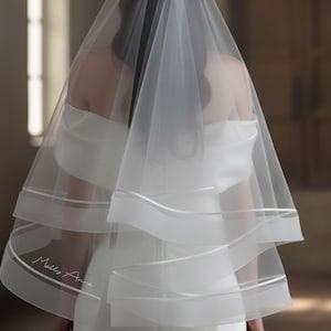Peut inclure: Un voile de mariée blanc transparent à plusieurs couches avec une bordure en ruban de satin. Le voile est drapé sur une robe de mariée blanche. Le nom "Molly Anne" est imprimé sur le voile.