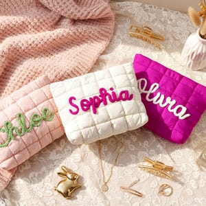 Op de afbeelding: Drie gewatteerde make-up tasjes in roze, wit en fuchsia, elk geborduurd met een naam: Chloe, Sophia en Olivia. De tasjes liggen op een kanten oppervlak met accessoires zoals haarclips, een gouden konijnenfiguur en een parfumflesje.