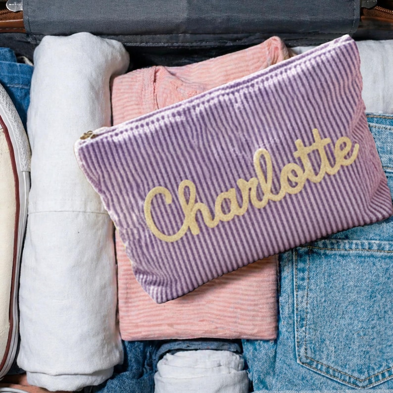 May include: A purple and white striped cosmetic bag with the name "Charlotte" embroidered in gold script. The bag is rectangular with a zipper closure and is surrounded by folded clothing and denim.