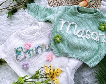 Könnte beinhalten: Zwei weiße Strickpullover mit gestickten Namen "Emma" und "Mason" in bunter Wolle. Die Pullover haben Blumenschmuck.