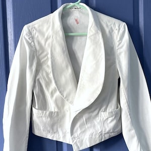 May include: A white, short-sleeved jacket with a shawl collar and two pockets. The jacket has a double-breasted closure and is made of a lightweight fabric.