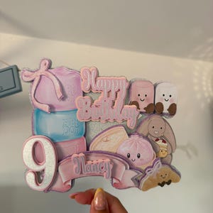 May include: A glittery cake topper featuring the text Happy Birthday and the name Nancy on a pink banner. The design includes a stack of colourful marshmallows, a large number 9, and cute illustrations of a bunny, a croissant, and a steamed bun.