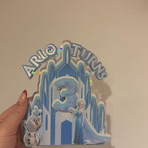 以下が含まれることがあります： 「Arlo Turns 3」の文字が入った青と白のケーキトッパー。映画「アナと雪の女王」のエルサとオラフのキャラクターが描かれたお城のデザイン。数字の3が大きく表示されています。