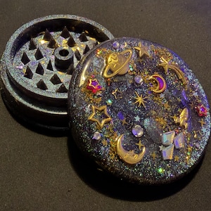 May include: A circular herb grinder with a dark, glittery resin finish. The lid features embedded gold-toned charms of a planet, crescent moon, stars, and a rocket ship, along with small clear crystals. The interior shows sharp, pyramid-shaped teeth.