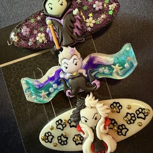 May include: Three hair clips featuring cartoon-style villain characters. The top clip is purple glitter with a horned figure. The middle clip has a blue and purple swirl pattern with a tentacled figure. The bottom clip is white with black paw prints.