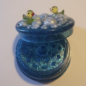 May include: A blue glittery container with a lid, featuring two small green duck figurines and clear bubble-shaped accents. The container is open, revealing a matching glittery interior.