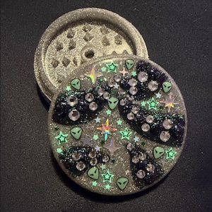 May include: A circular herb grinder with a silver glitter finish. The top lid features a space-themed design with dark purple glitter clouds, small light green alien head charms, iridescent star-shaped sequins, and clear round rhinestones.