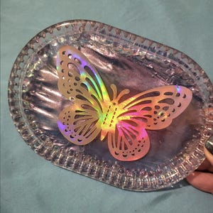 May include: An oval dish with a textured edge and an iridescent sheen. A translucent butterfly with rainbow wings sits in the centre. The butterfly has a cut-out pattern on its wings.