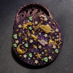May include: A circular, two-piece resin herb grinder in deep purple with a glittery finish. The surface is embedded with gold-toned charms including a rocket, a planet with rings, a crescent moon, and stars, alongside small green alien face icons.