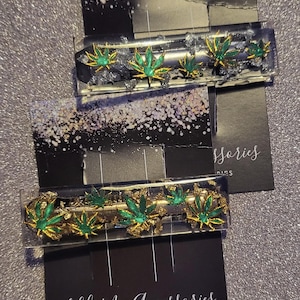 May include: Two rectangular hair clips with green and gold marijuana leaf designs. The clips are set in clear resin and are displayed on black cards with the text "LillyMaiAccessories". The background is a sparkly silver surface.