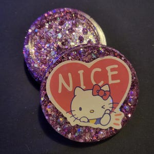 May include: Two round, purple glitter resin coasters. One coaster has a Hello Kitty graphic inside a red heart with the word "NICE". The other coaster is covered in purple glitter.