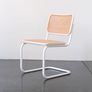 Puede incluir: Una silla moderna con estructura de metal blanco y asiento y respaldo de mimbre tejido. La silla tiene un diseño minimalista, con una base curva y un respaldo rectangular. La silla está colocada contra una pared blanca y un suelo gris oscuro.