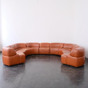 May include: A large, curved, sectional sofa in a warm, orange-brown leather. The sofa is composed of multiple sections, creating a rounded shape. The design is modern and minimalist, with a focus on comfort and style.