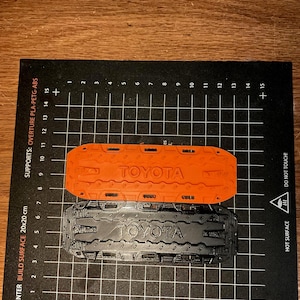 May include: Two 3D-printed Toyota accessories, one orange and one black, rest on a black surface with a grid pattern. The Toyota logo is visible on each item. The surface has text indicating it is a 3D printer build surface.