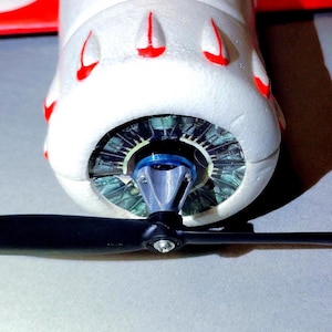 May include: Close-up of a model airplane's nose. The white foam body has red accents and a detailed engine design. A black propeller is attached to the front, with a blue ring around the engine's center.