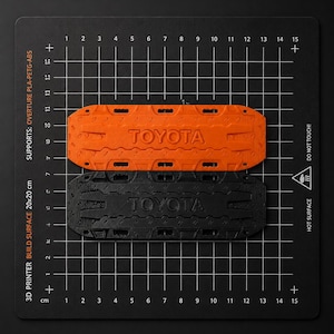 May include: Two rectangular recovery boards, one orange and one black, each featuring the embossed text TOYOTA. These items are positioned on a black 3D printer build surface measuring 20x20 cm, which includes a grid and safety warnings.