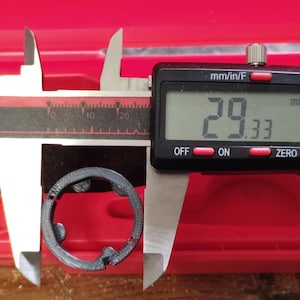 May include: A digital caliper measuring a dark gray, circular object. The digital display reads 29.33 mm, approximately 1.15 inches. The caliper is set against a red background, with the object resting on a wooden surface.