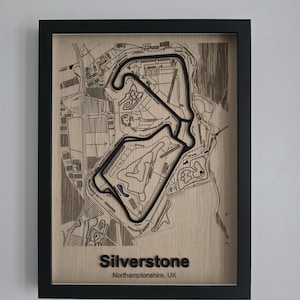 May include: A framed wooden wall art piece featuring a laser-engraved map of the Silverstone race circuit in Northamptonshire, UK. The track is highlighted in black, set against a light wood background with text reading Silverstone Northamptonshire, UK.
