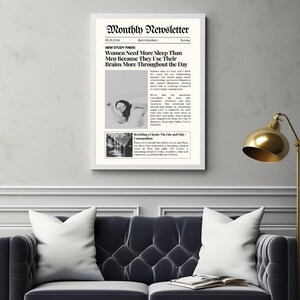 May include: A framed print titled "Monthly Newsletter" with text and images. The headline reads "Women Need More Sleep Than Men Because They Use Their Brains More Throughout the Day." The print is displayed above a dark gray sofa with white pillows.