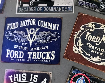Könnte beinhalten: Eine Sammlung von Vintage-Metallschildern mit Ford-Lastwagen, -Autos und -Logos. Die Schilder sind in verschiedenen Farben, darunter Rot, Blau, Weiß und Schwarz. Die Schilder zeigen Texte wie "F-Series Trucks", "Ford Motor Company", "Ford Trucks", "This is a Ford Garage" und "Shelby GT 350".