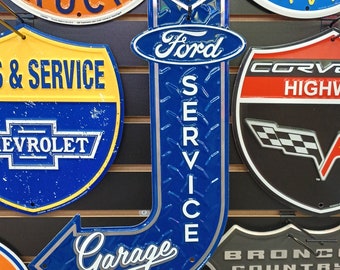 Könnte beinhalten: Eine Sammlung von Vintage-Metallschilden mit verschiedenen Automarken, darunter GMC Trucks, Chevrolet, Ford, Corvette, Pontiac und Bronco. Die Schilder sind in verschiedenen Farben lackiert, darunter Blau, Rot, Gelb und Weiß. Die Schilder werden an einer Holzwand angezeigt.