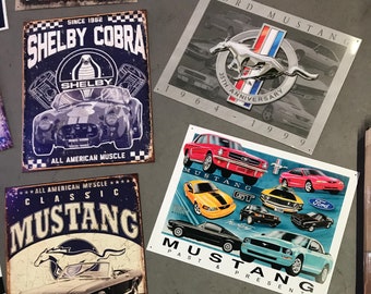Könnte beinhalten: Eine Sammlung von Vintage-Blechschildern mit verschiedenen Ford Mustang-Modellen. Die Schilder sind in verschiedenen Farben gehalten und zeigen klassische Mustang-Logos und -Texte, darunter "Shelby Cobra", "Classic Mustang", "Mustang GT" und "Americanbred Since 64".