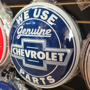 Puede incluir: Un letrero redondo azul y blanco de Chevrolet con el texto "WE USE Genuine CHEVROLET PARTS". El letrero se exhibe con otros letreros similares, todos envueltos en pl&aacute;stico.