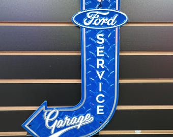 Könnte beinhalten: Blaues und silbernes Metallschild mit einem Diamantenmuster. Das Schild ist in Form eines nach rechts zeigenden Pfeils geformt und trägt die Aufschrift "Ford Service Garage" in weißen Buchstaben.