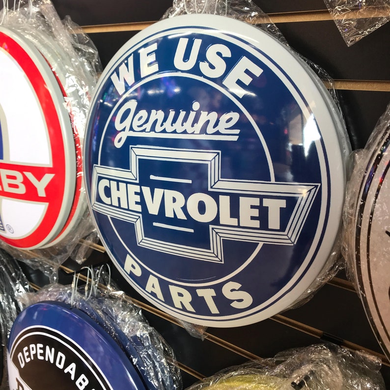 Puede incluir: Un letrero redondo azul y blanco de Chevrolet con el texto "WE USE Genuine CHEVROLET PARTS". El letrero se exhibe con otros letreros similares, todos envueltos en pl&aacute;stico.