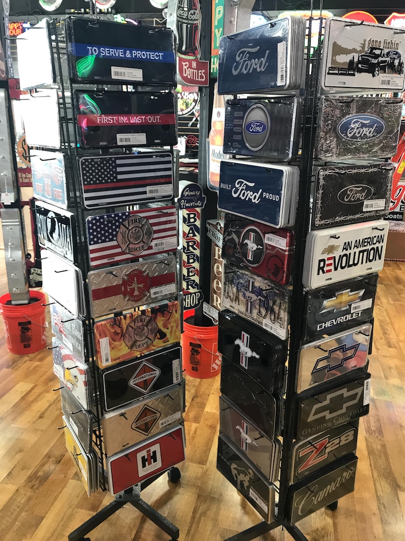 May include: A metal display rack with various license plate-style metal signs. The signs feature different logos and slogans, including "To Serve and Protect", "First In, Last Out", "Built Ford Proud", "An American Revolution", and "Chevrolet".