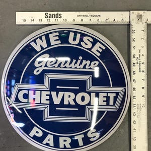 Puede incluir: Un letrero redondo de metal blanco y azul con el texto "WE USE Genuine CHEVROLET PARTS" en letras blancas sobre fondo azul.