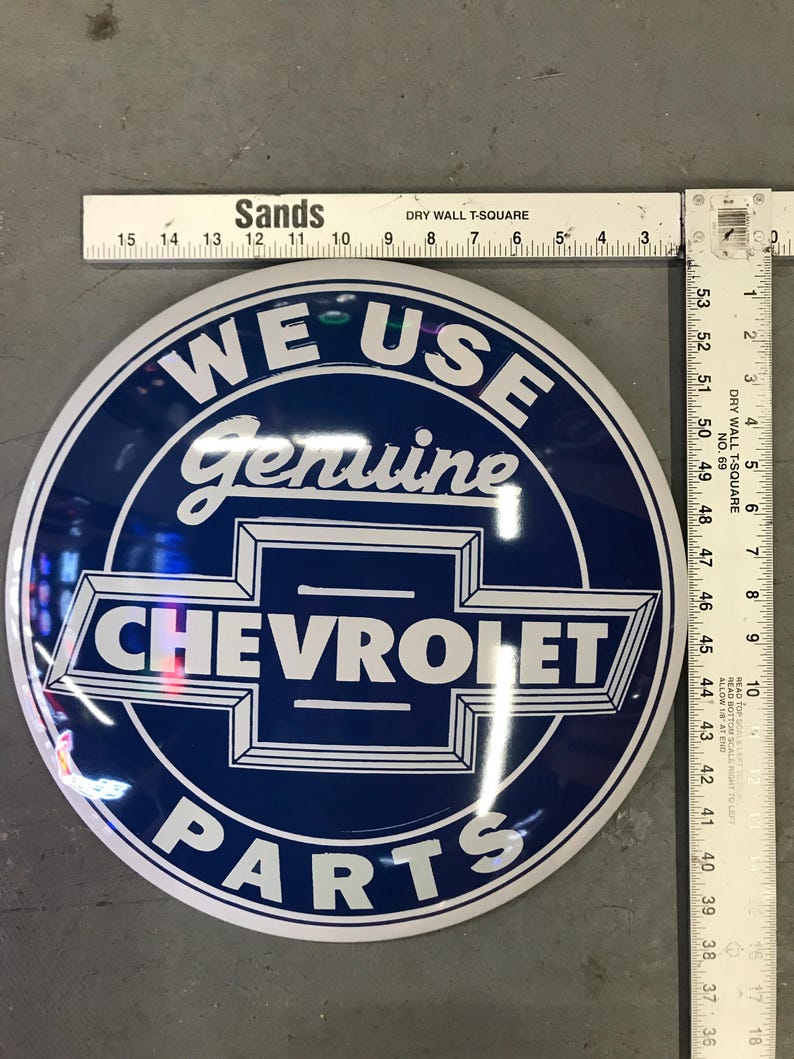 Puede incluir: Un letrero redondo de metal blanco y azul con el texto "WE USE Genuine CHEVROLET PARTS" en letras blancas sobre fondo azul.