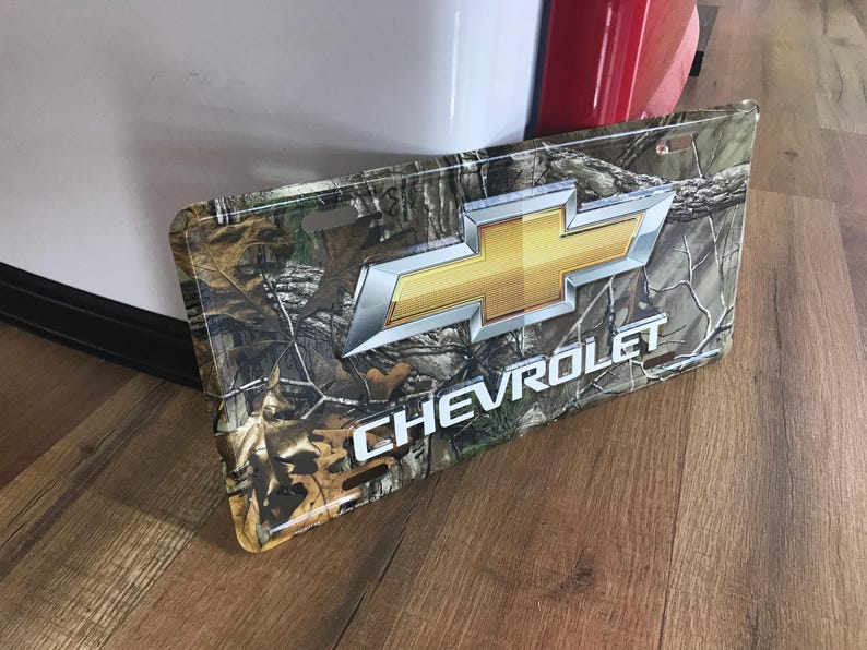 May include: A metal license plate featuring a camouflage pattern with a gold Chevrolet logo and the word "CHEVROLET" in white lettering.